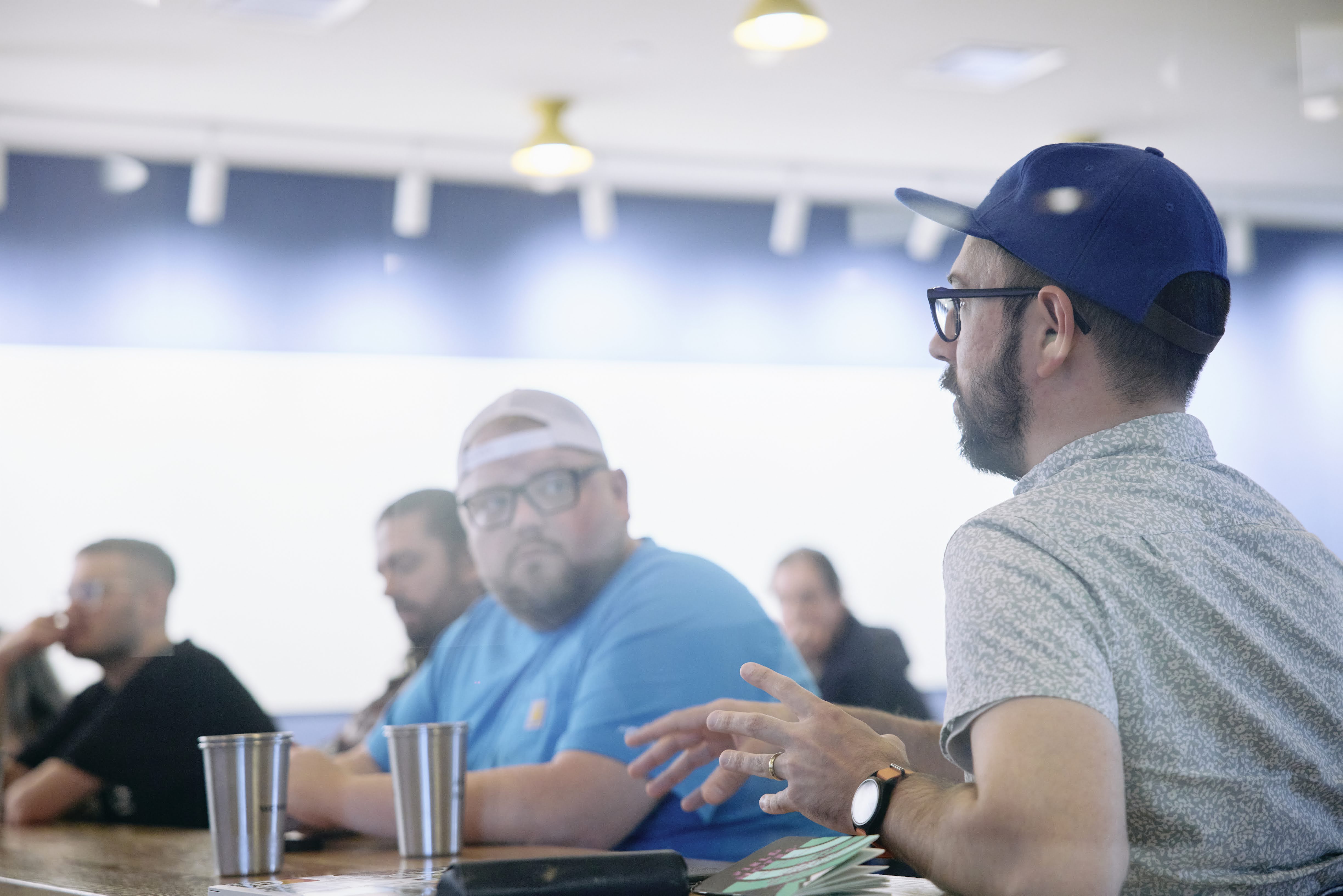 Drew in the foreground gesturing and speaking with a room of Savas colleagues.